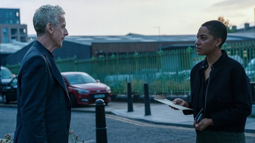 Peter Capaldi and Cush Jumbo stand facing each other on a quiet road