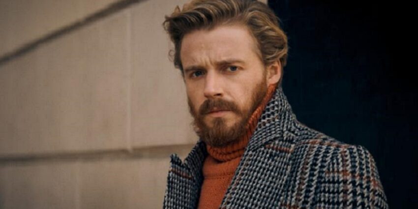 Bearded actor Jack Lowden stands outside in a tweed coat and brown polo-neck sweater