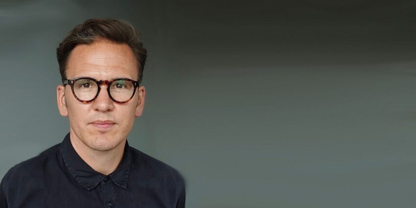 Close-up of Karl Warner, in dark shirt with glasses, against a dark background