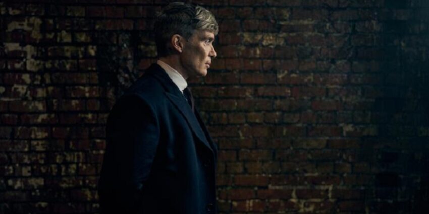 Dark photo of Cillian Muphy as Tommy Shelby in Peaky Blinders, standing against a brick wall