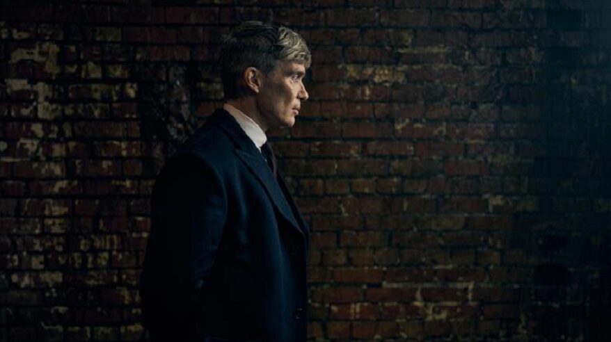 Dark photo of Cillian Muphy as Tommy Shelby in Peaky Blinders, standing against a brick wall