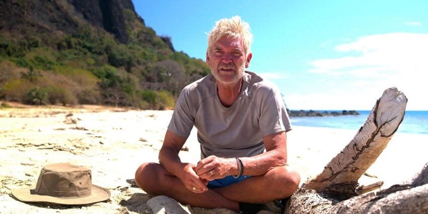 Phillip Schofield sits unshaven on a remote beach in a grey t-shirt