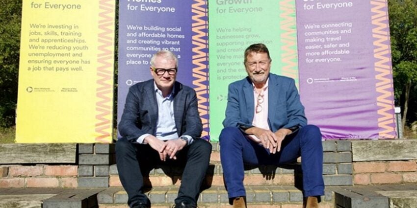 Richard Parker and Steven Knight sitting outdoors on stone steps with coloured banners of writing behind them