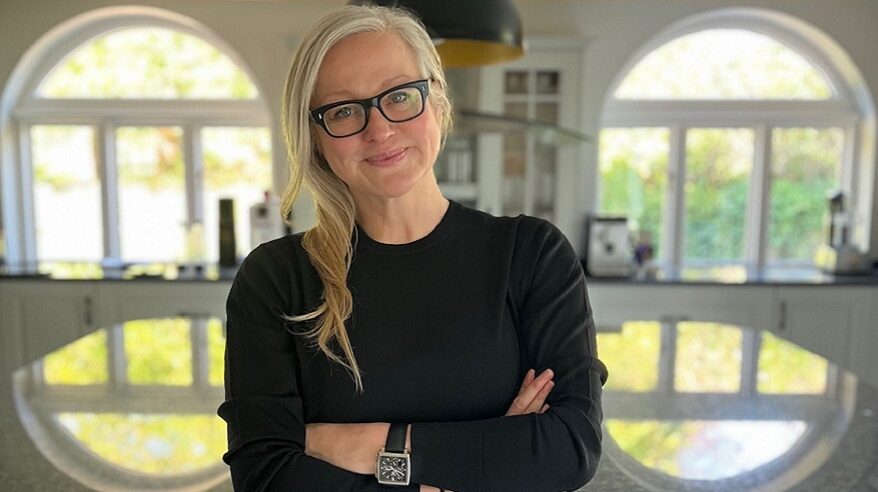 Produce Nickie Sault, wearing a black sweater and wristwatch, standing with arms folded in a room with windows behind