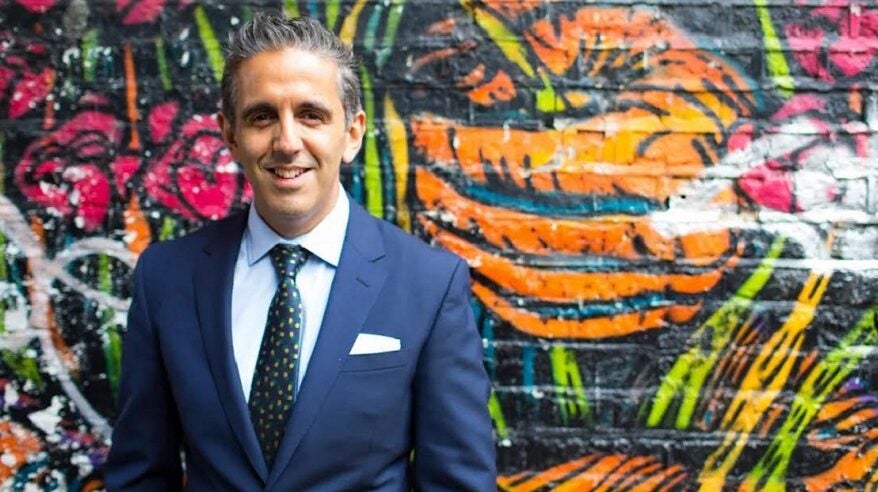 Barrister and writer Imran Mahmood in blue suit and green tie standing against a brightly-painted outside wall