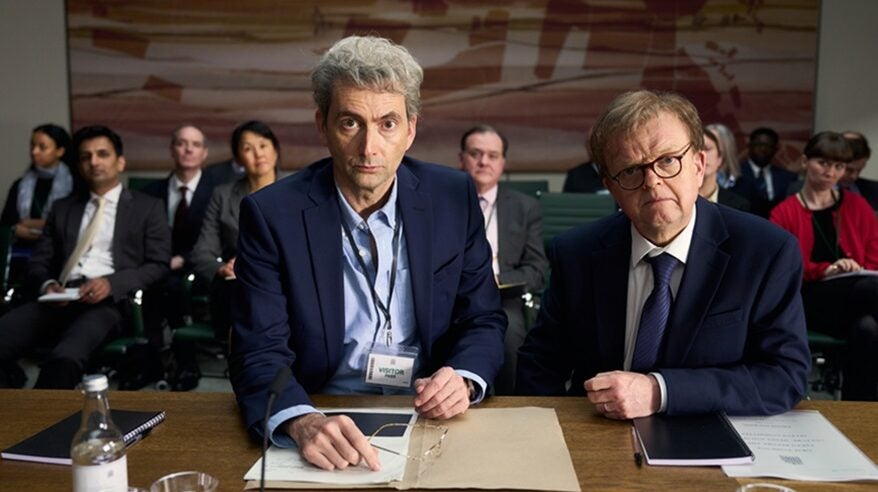 Actors David Tennant and Toby Jones in character in new ITV drama The Hack, seated next to each other at a desk with people behind