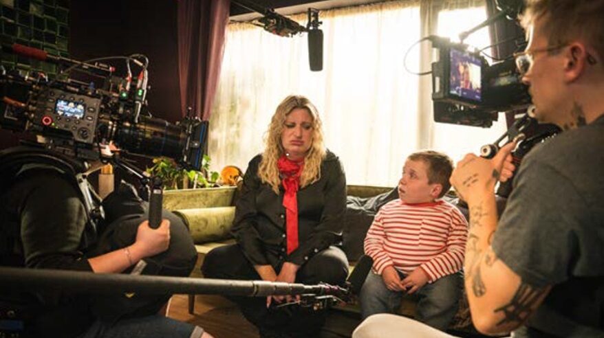 Actors Daisy May Cooper and Lenny Rush sitting on a sofa with a camera mic boom in front of them