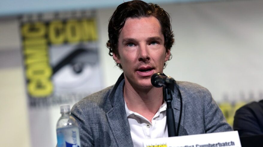 Close-up of actor Benedict Cumberbatch in grey jacket sitting at a table with a microphone