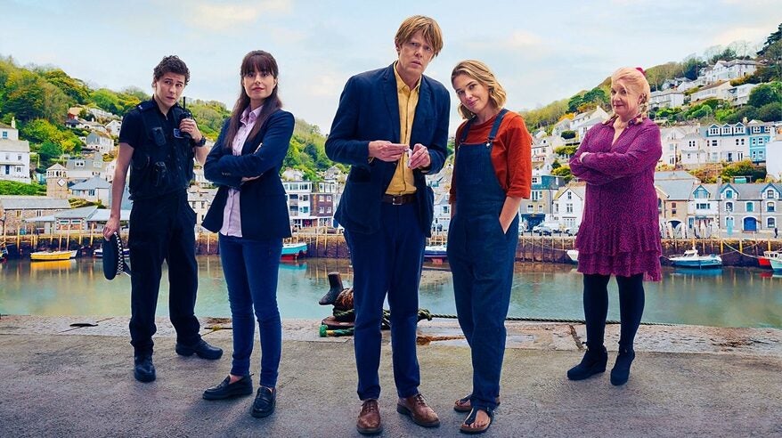 The cast of BBC drama Beyond Paradise standing together on a harbour wall with a coastal village backdrop