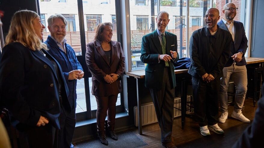 Group of people standing against a window with HRH The Duke of Edinburgh in the middle, wearing a suit and smiling