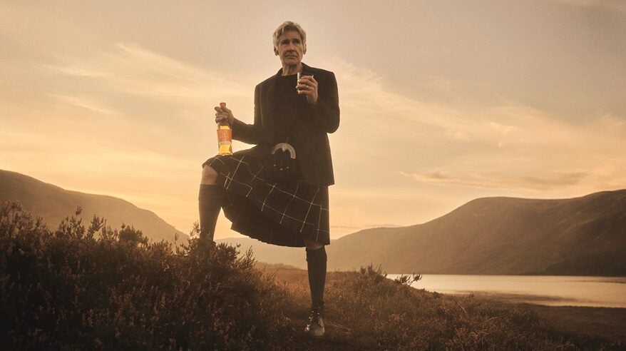 Actor Harrison Ford stands on a Scottish mountain wearing a kilt and holding a bottle of whisky