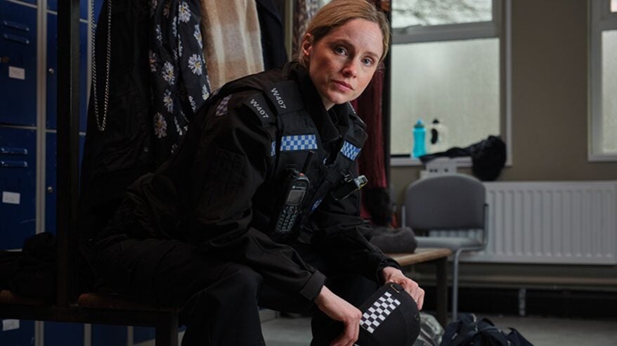 Actor Sophie Rundle in character wearing police uniform in ITV drama After the Flood, seated with coats and chairs behind her