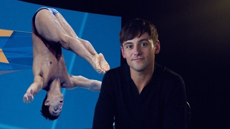 British Olympic diver Tom Daley in a black shirt in front of a photo of him diving