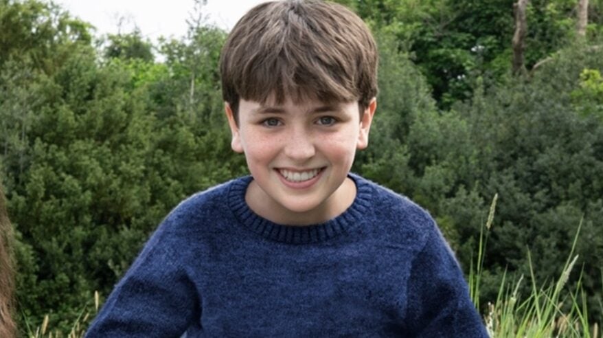 Young actor Dominic McLaughlin sitting in long grass with trees behind, wearing a navy sweater