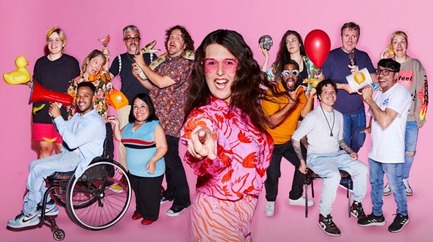 Comedian Rosie Jones wearing a pink patterned top and orange glasses with a group of people behind her against a pink background