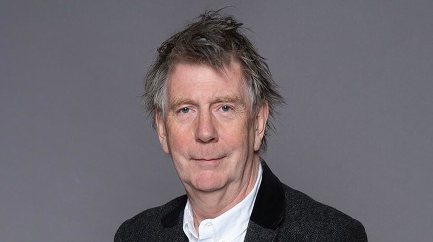 Headshot of ITV's John Whiston, wearing a white, open-necked shirt and black jacket