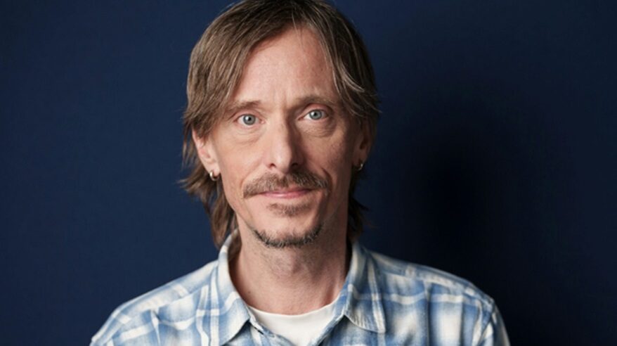 Actor Mackenzie Crook in checked shirt with white tee underneath, against a dark background