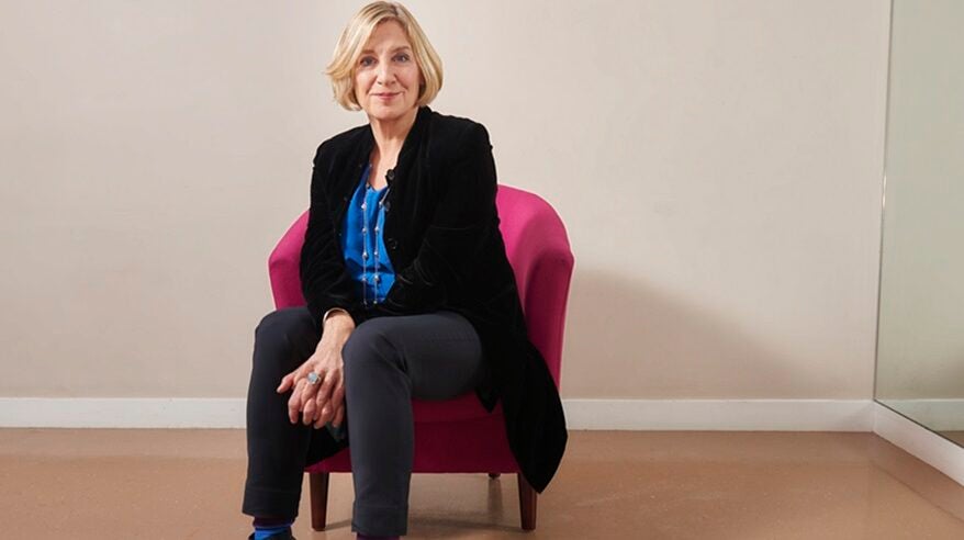 The late comedian, actor and writer Victoria Wood seated on a pink chair, wearing a black jacket over a blue shirt
