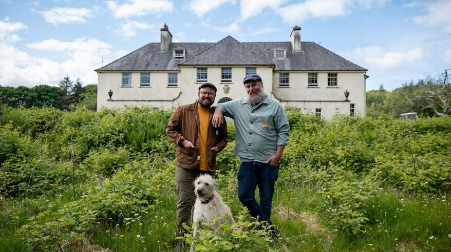 Two men wearing caps stand on an overgrown lawn in front of a derelict mansion, with a white lurcher dog at their feet