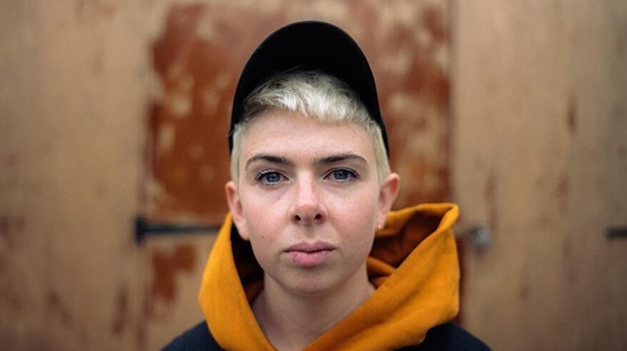 Director Molly Manning Walker, with short blonde hair under a black cap, wearing orange hoodie