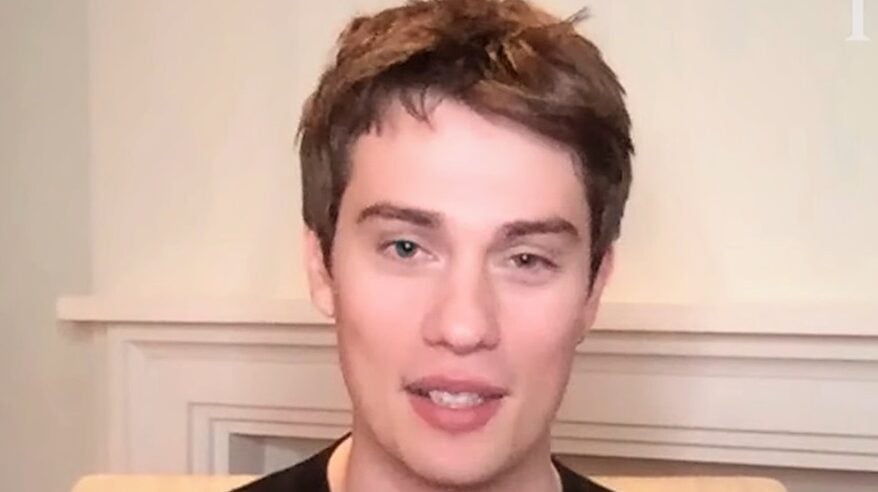 Headshot of actor Nicholas Galitzine wearing black top, against a pale wall and fireplace