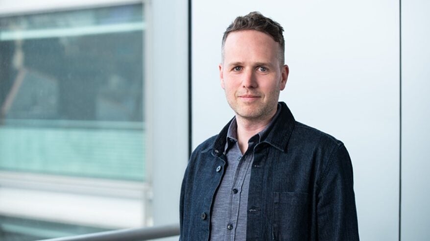 Film executive Ollie Madden pictured against a white wall, wearing a grey shirt and navy jacket