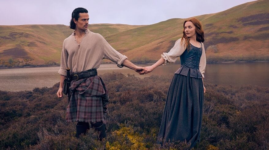 Two actors in Scottish period costume stand on a moor hand in hand, from Starz drama Outlander: Blood of my Blood