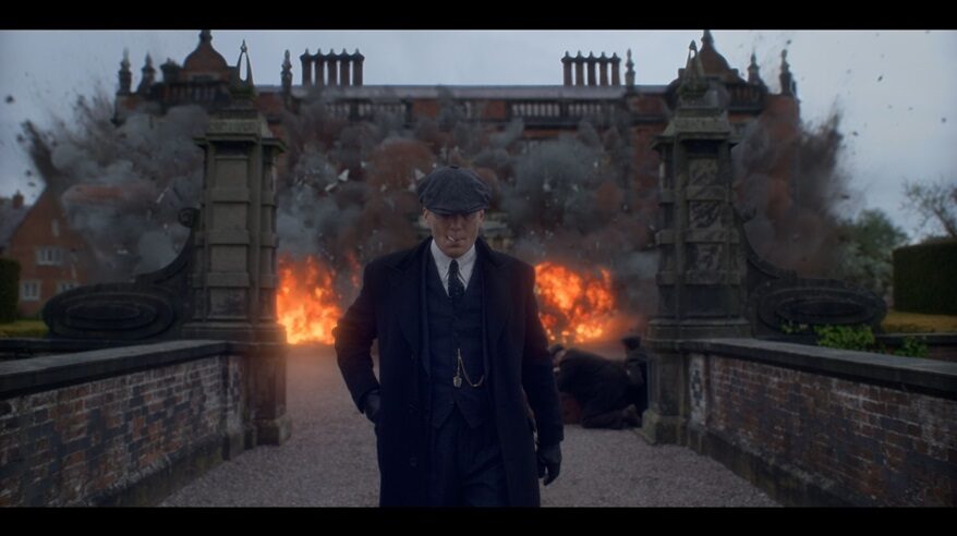 Cillian Murphy in Peaky Blinders, wearing a shirt, tie, coat and cap, walking away from a house exploding into flames behind him