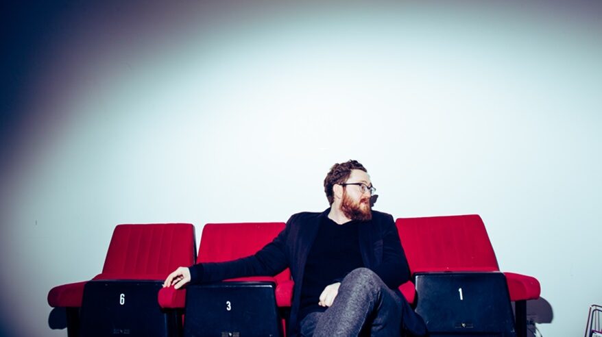 Red-haired, bearded man in glasses on red cinema seats looking to his left