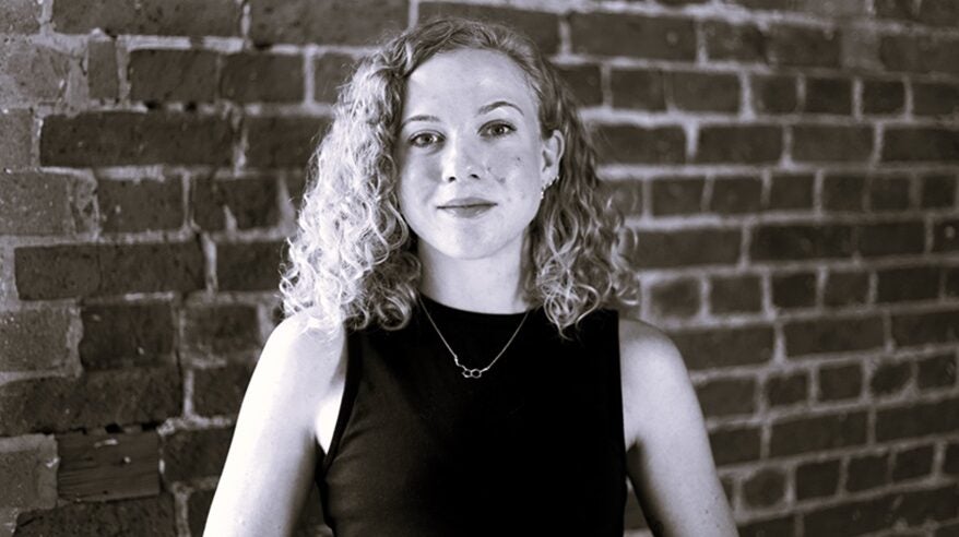 Black and white image of a lady with long curly fair hair in a black top with brick wall in background