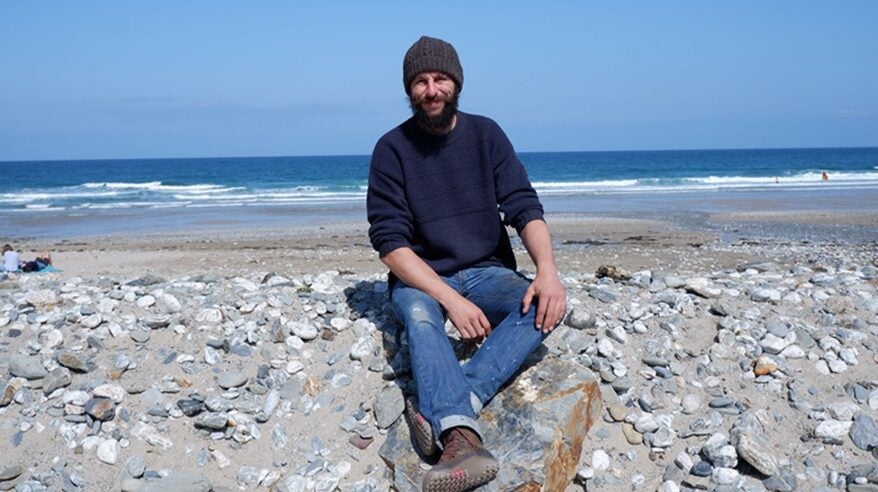 Bearded man in hat, sweater, jeans and boots sits on pebbly beach with sea and sky behind