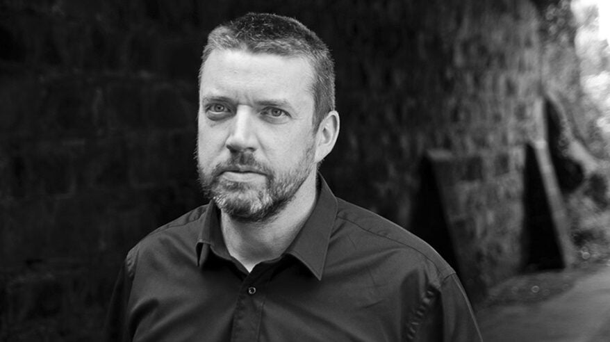 Author Liam McIlvanney in black and white photo wearing a dark shirt standing in front of an outside wall