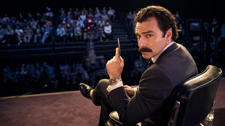 A suited man with a moustache turns on this chair to face behind him, away from a studio audience, in an image from drama Rivals on Disney+