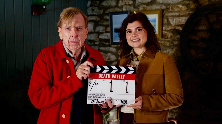 A fair-haired man in a red jacket and a dark-haired lady in a brown jacket jointly hold a film clapperboard
