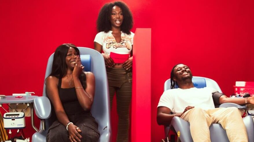 Two people sit in blood donation chairs separated by a screen, with a lady standing behind them, all against a red background