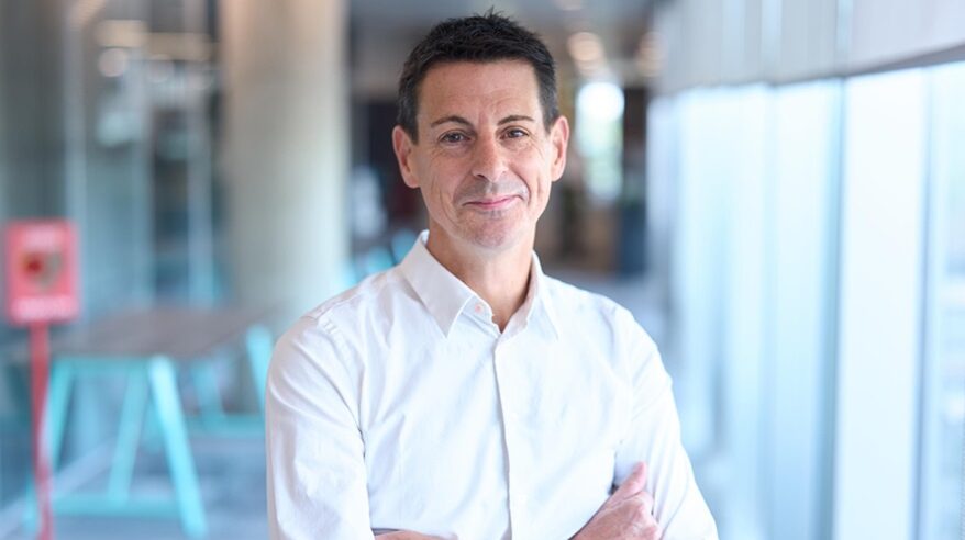 Dark-haired man wearing white shirt stands with arms folded, smiling to camera