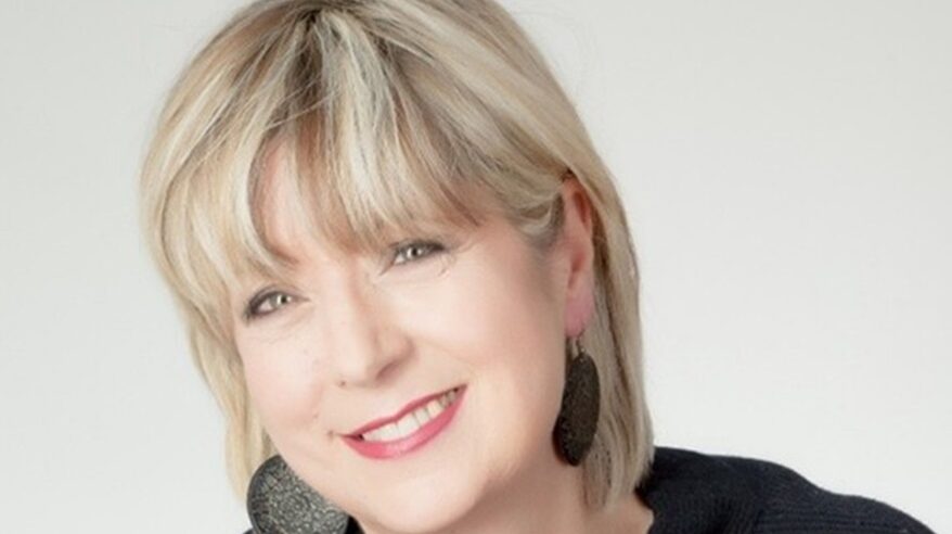 A blonde lady with large disc earrings and black top smiles to camera