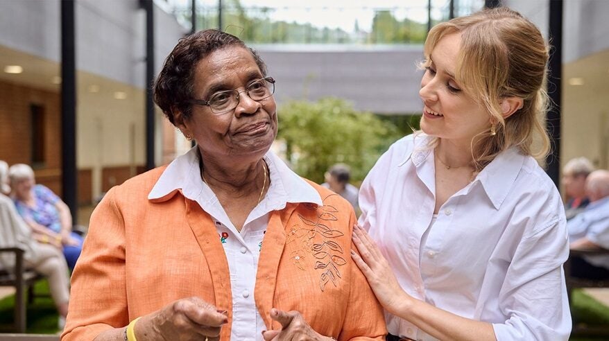 An older lady in a peach jacket and glasses stands next to a younger lady in a white shirt
