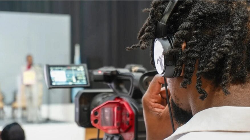 A young person with short black hair seen from behind operating a TV camera and wearing headphones