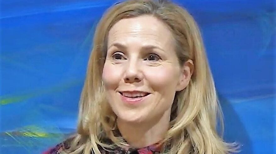 Blonde lady sits smiling in patterned top against a royal blue background