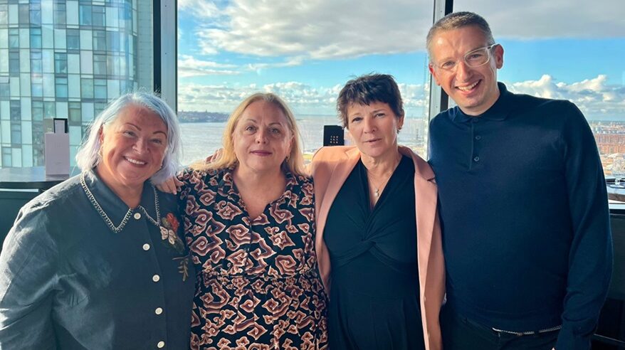 Three women and one man stand with arms linked behind their backs in front of a large window with blue sky and clouds behind
