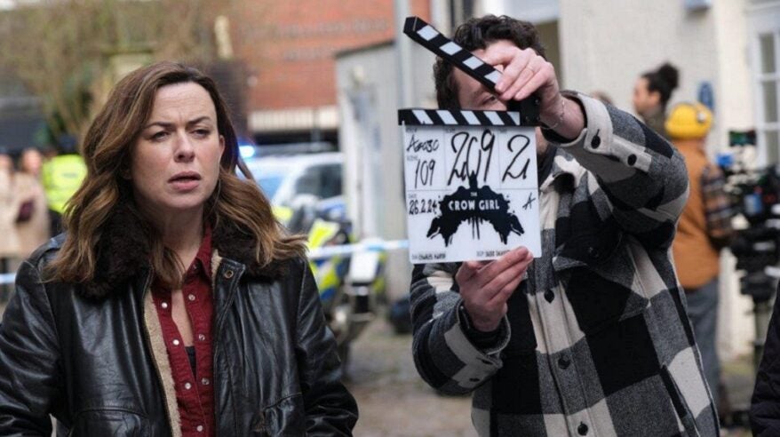 A lady with shoulder-length brown hair (actor Eve Myles) in a street with a film clapperboard in front of her