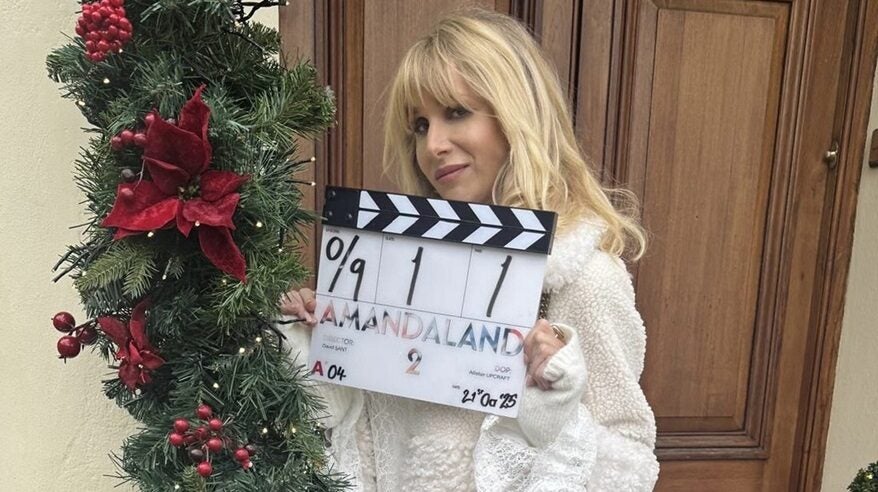 A blonde woman in a white jacket stands in front of a wooden door, decorated with Christmas greenery and red ribbon, holding a clapperboard for the start of filming on Amandaland Christmas special