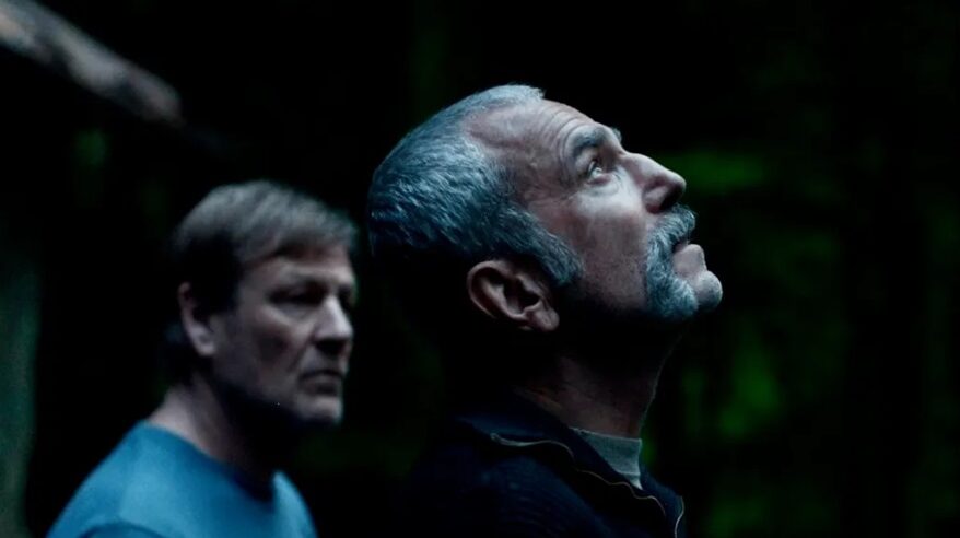 A bearded, silver-haired man tilts his head back to look up at the sky, with a dark background and a man standing behind him, in an image from the film Anemone