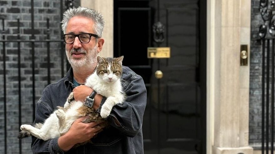Man wearing glasses with silver hair and beard holds a grey and white cat in his arms outside No 10 Downing Street front door