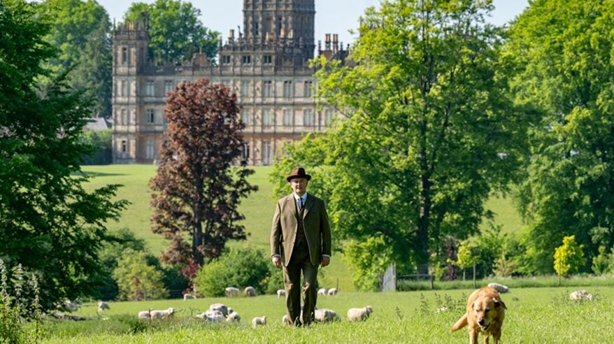 A man in 1930s outdoor clothing stands with sheep and a dog in front of a grand stately home and fields