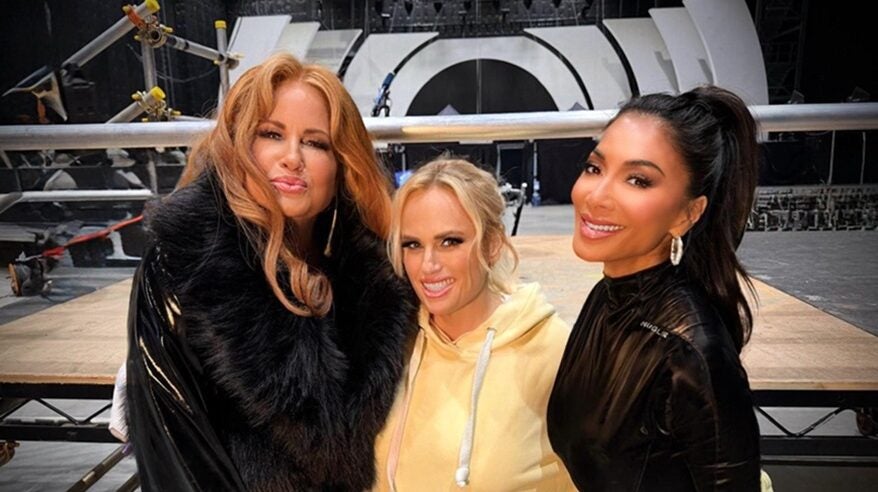 Three glamorous ladies stand together, smiling to camera, with a large rigged arena behind them
