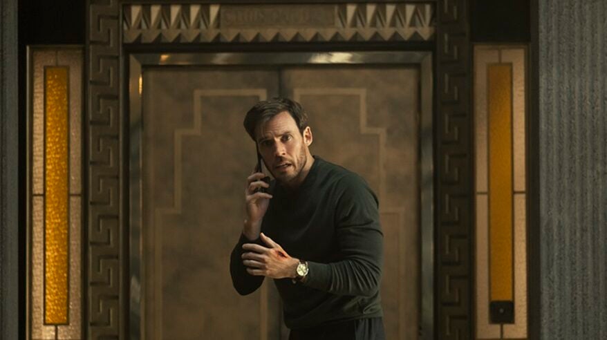 A young bearded man in a dark green sweater stands with his phone to his ear in front of a bronze interior wall with an Art Deco pattern