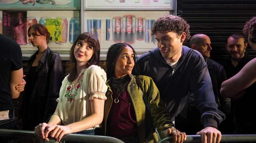 Two young women stand at a barrier smiling at a curly-haired young man next to them
