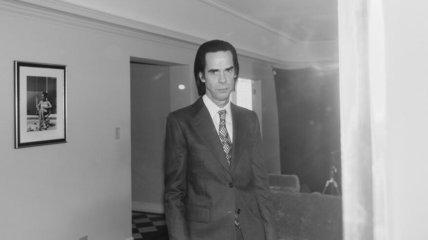 Black and white shot of a tall man, musician Nick Cave, in suit and tie with swept back dark hair standing in a living room